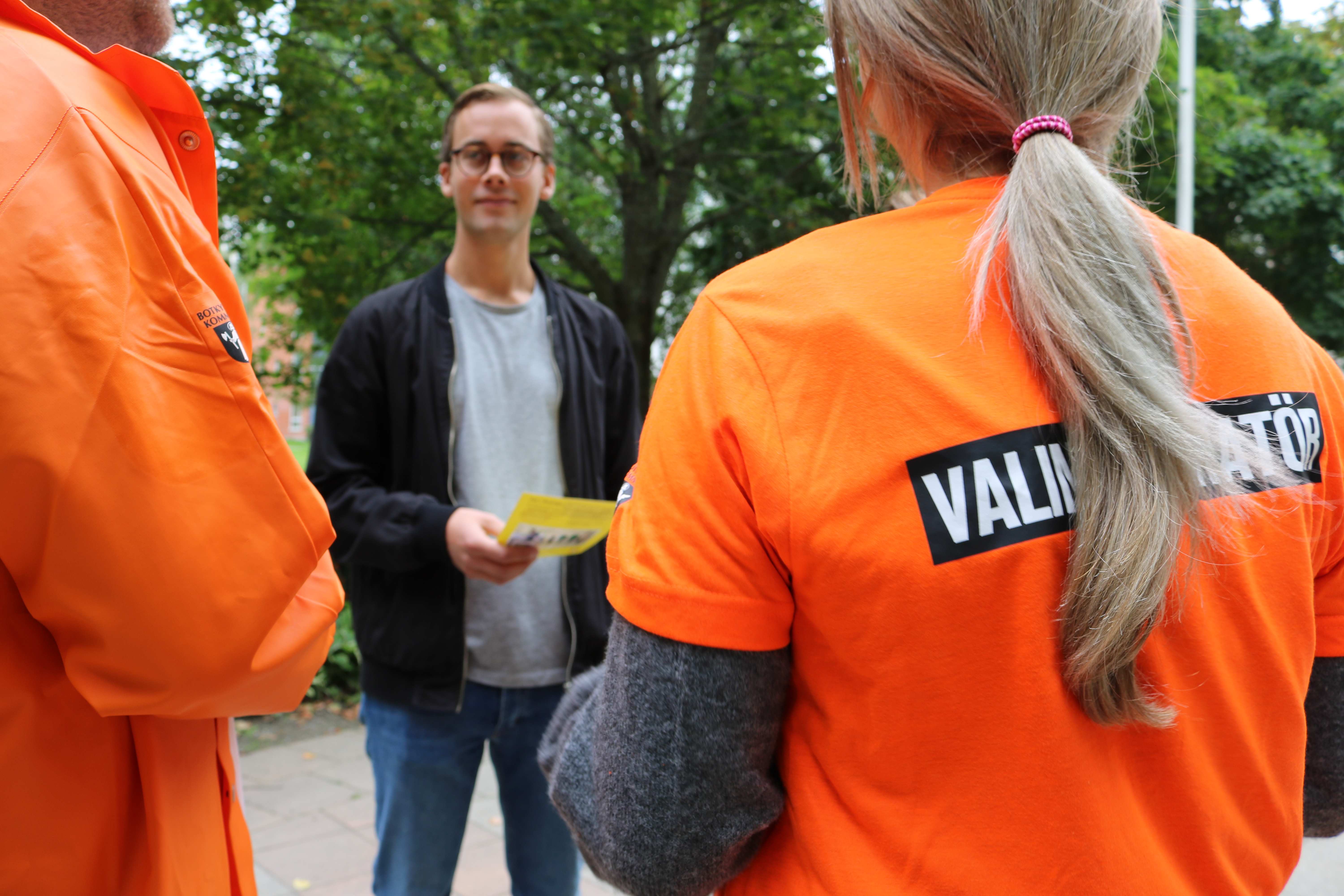 Två valinformatörer syns bakifrån. På ryggen på deras orangea kläder syns texten "Valinformatör"