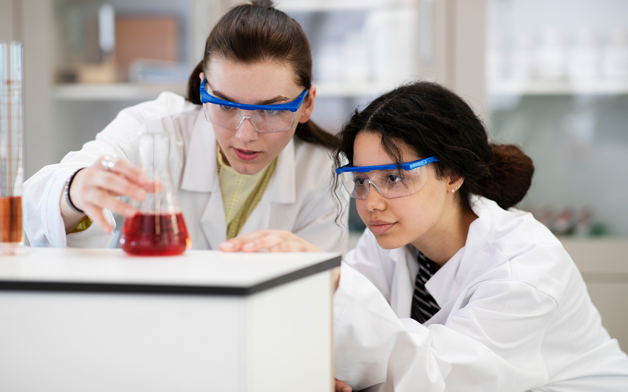 Två elever med skyddsglasögon och vita rockar tittar på en röd vätska i ett provglas.