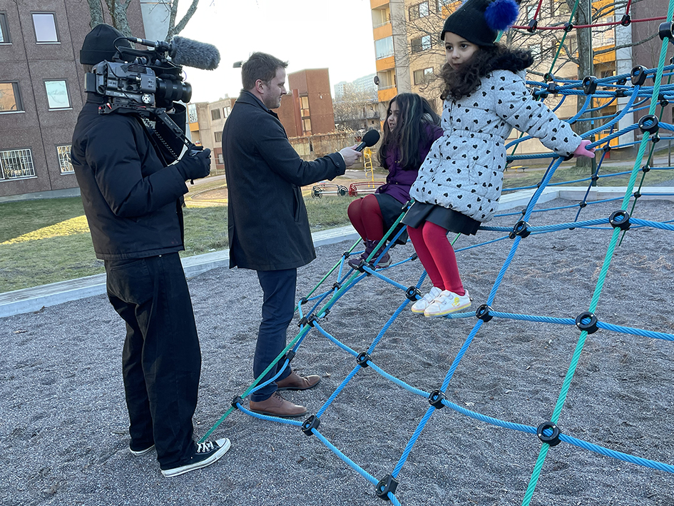 Två vuxna och två barn i en lekpark. En vuxen filmar med filmkamera, en vuxen håller fram en mikrofon till barnen. Barnen sitter i en klätterställning. Solen skiner.