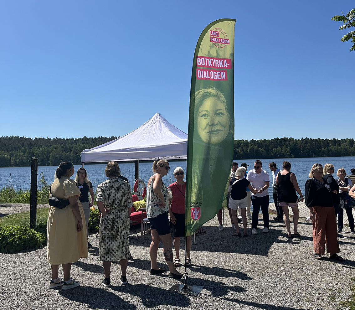 Solen skiner i en park. I bakgrunden blå himmel och blått vatten, i förgrunden flertalet människor som minglar och i mitten en strandflagga med ett kvinnoansikte och texten Botkyrkadialogen.