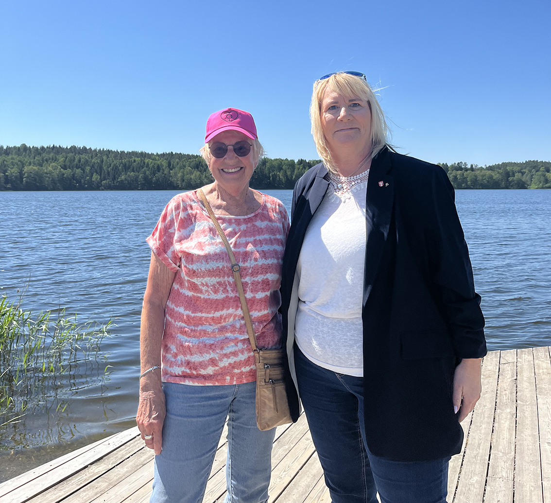 Två kvinnor står tillsammans på en brygga. Solen skiner och himlen i bakgrunden är klarblå.