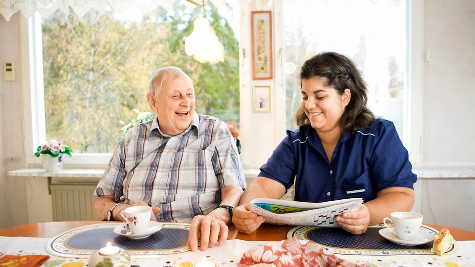 En äldre man och en kvinna vid ett köksbord med kaffekoppar och en dagstidning.