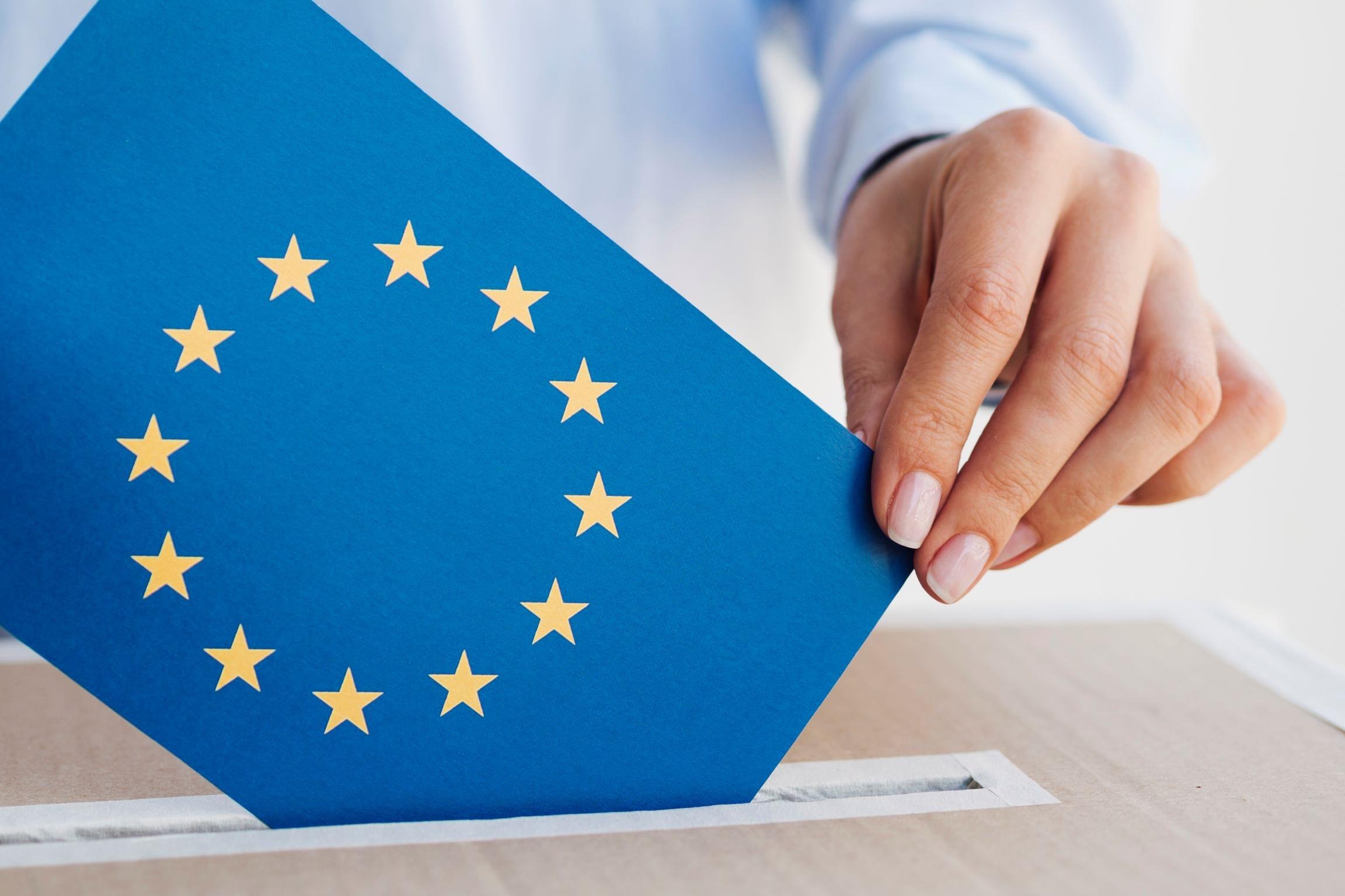 En hand som lägger ett EU-flaggsfärgat röstkuvert i en röstlåda.