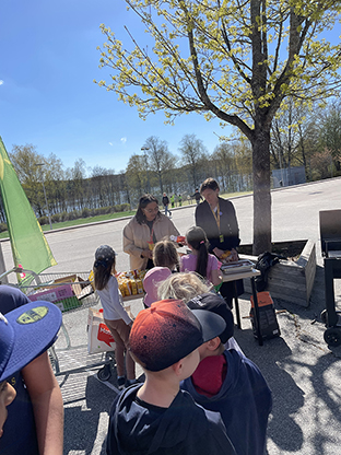 En solig dag med klarblå himmel och en spirande grönt träd i bakgrunden. I förgrunden barn med ryggarna mot kameran köar till en grill för att få korv och dricka.