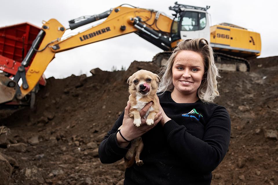Anna Jansson medarbetare på återvinningsföretaget Hummeltorp bär på lilla hunden Pelle. I bakgrunden syns företagets största grävskopa som Anna brukar köra.