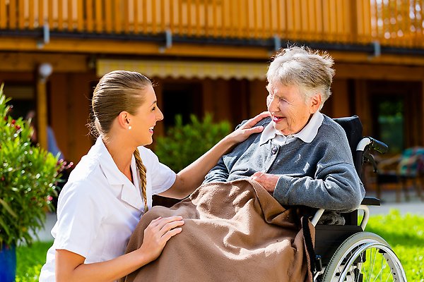 Vårdpersonal, en ung kvinna skrattar med en äldre dam i rullstol, de är utomhus i solen