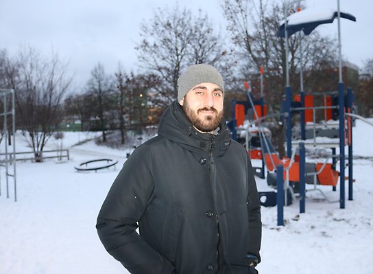 Ung man med skägg och vinterkläder står i en snöig park med lekutrustning i bakgrunden. Han har händerna i fickorna och ler mot kameran.