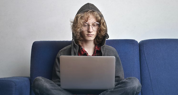 Kille med luvtröja, lockigt hår och glasögon sitter i en soffa med en grå bärbar dator i knäet.